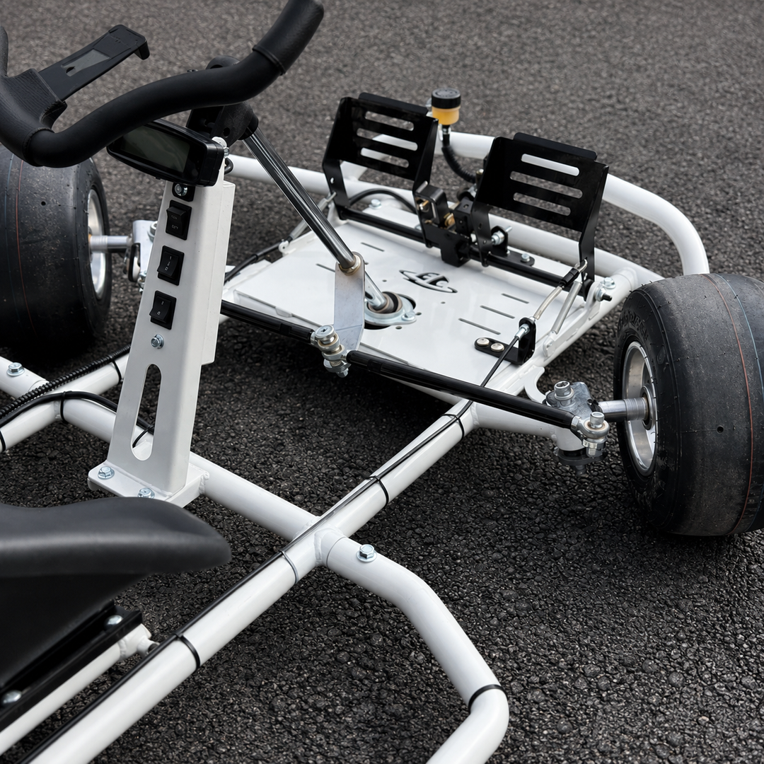 A close-up of a white go-kart chassis with black tires, black seat, and black footrests, photographed on dark asphalt pavement.