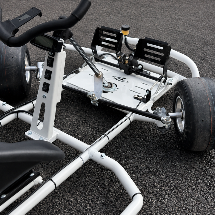 A close-up of a white go-kart chassis with black tires, black seat, and black footrests, photographed on dark asphalt pavement.
