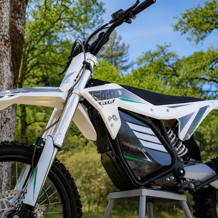 A white and black electric dirt bike with green accents, shown outdoors on a metal stand with trees and blue sky in the background.