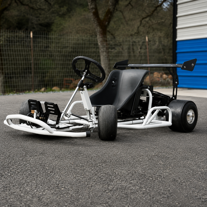 A go-kart with a black seat, black steering wheel, and white tubular frame, parked on an asphalt surface with a fence and trees in the background.