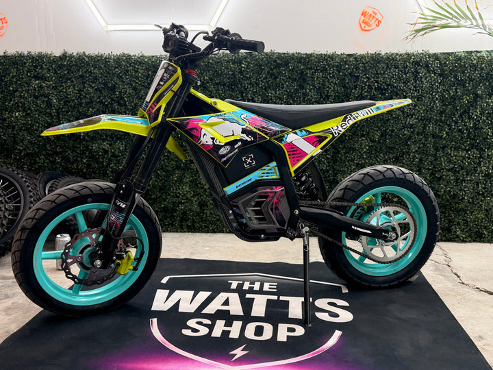 A small electric dirt bike with a vibrant multicolor graphic wrap, standing on its kickstand on a black mat printed with 'THE WATTS SHOP' logo. The bike features lime green, pink, and black graphics with turquoise accents on the rims.