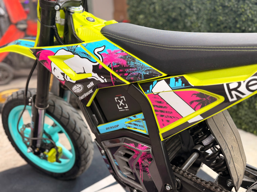 Close-up of a small electric dirt bike with a bright neon yellow frame, black seat, black battery housing, and colorful graphic decals in pink, blue, white, and black. The bike is photographed outdoors on a paved surface with a hedge in the background.