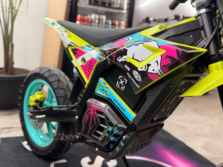 A close-up of a custom electric dirt bike with a vibrant lime green, black, and pink graphic livery. The bike features a black seat, a black carbon-fiber-look body panel with pink, blue, and white graphics, and a turquoise wheel with a black tire.