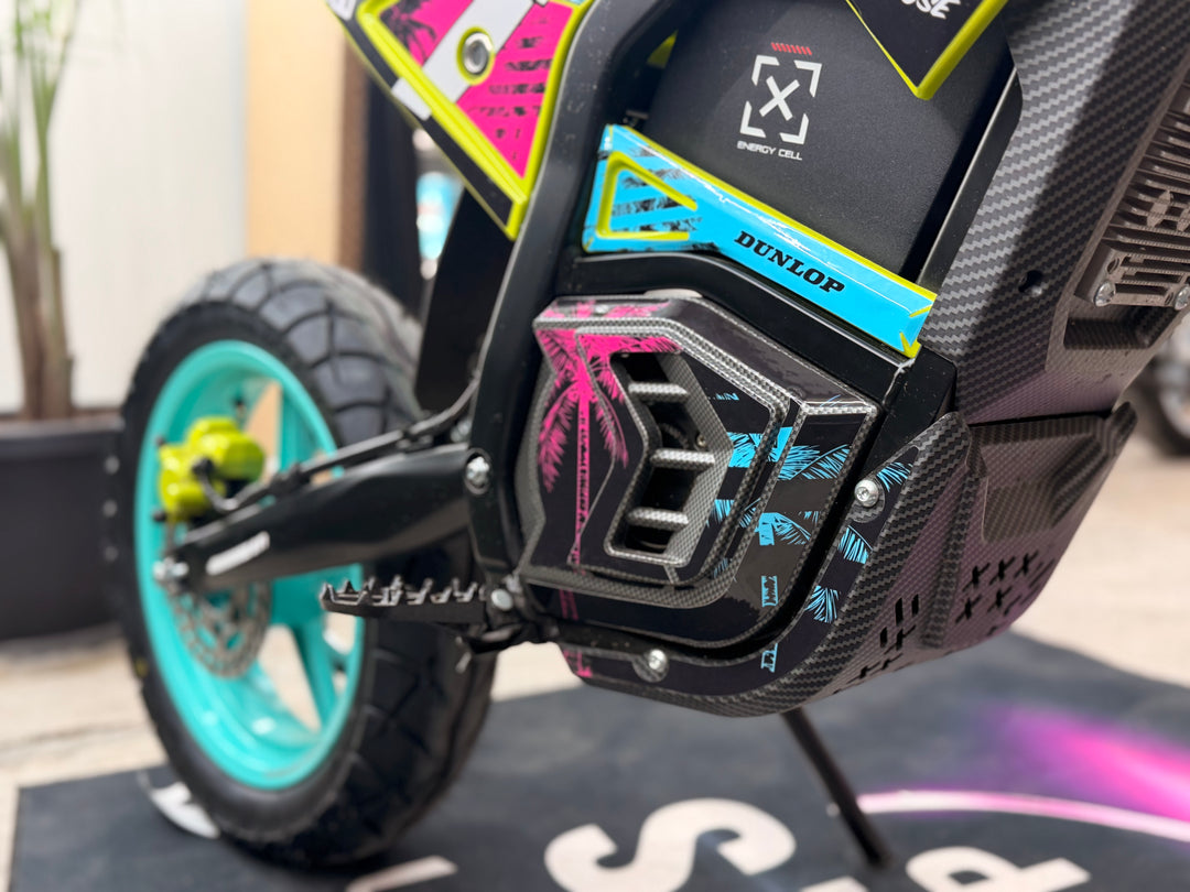 Close-up of a carbon fiber motorcycle engine cover with pink and blue graphics, featuring a visible Dunlop logo, alongside a turquoise wheel and black tire.