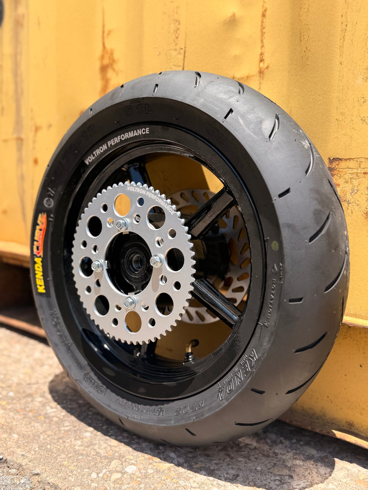 A black motorcycle tire mounted on a black wheel with a silver disc brake and sprocket, photographed outdoors against a yellow metal container.